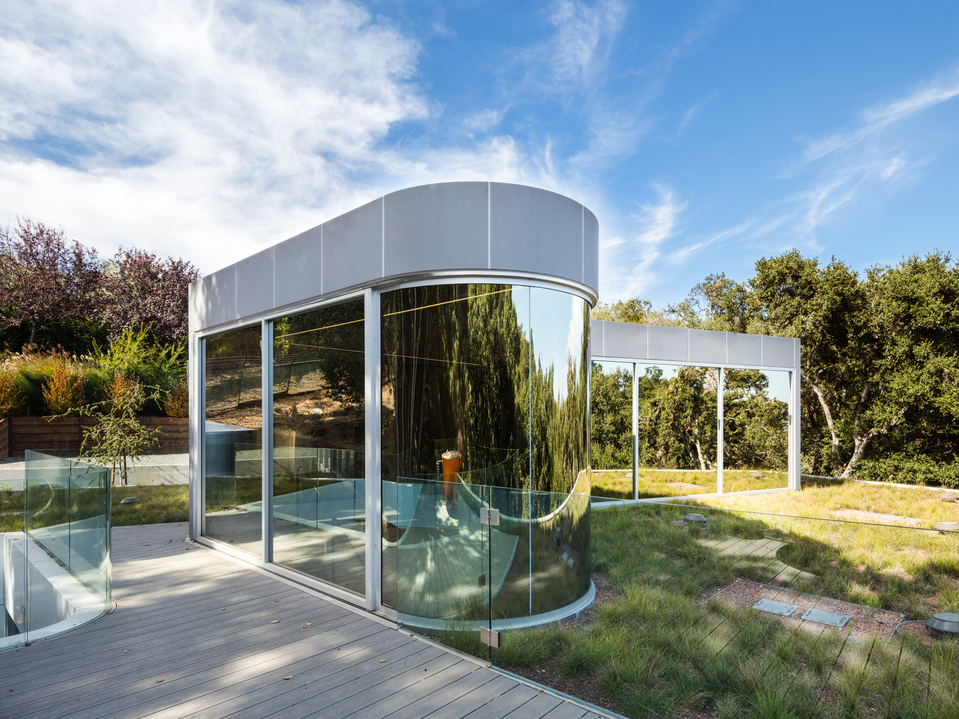 房子頂層圓錐玻璃量體宛如雕塑藝術品，右側牆面以鏡面玻璃映照周圍綠樹和藍天。